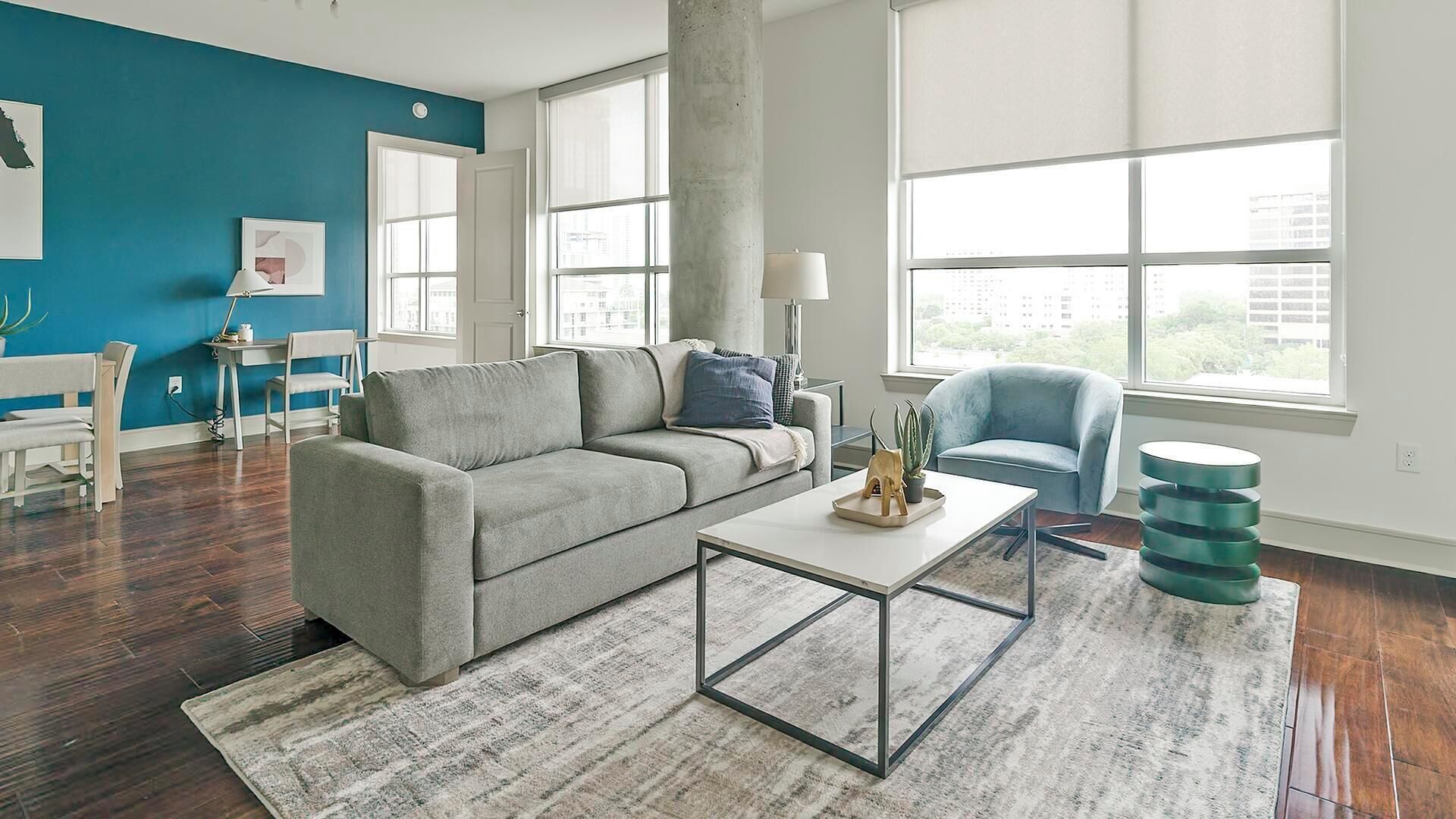 Uma sala de estar moderna com um sofá cinzento, uma mesa de centro e uma poltrona azul. Há grandes janelas que proporcionam luz natural e vista para a cidade.