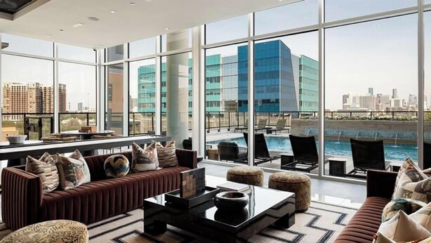 Une chambre d’hôtel moderne dotée de grandes fenêtres, d’un coin salon avec des canapés et des poufs, et offrant une vue sur les toits de la ville et sur le fleuve.