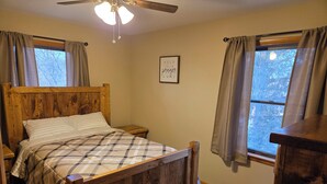 Ein Bett mit Holzbettgestell und karierter Bettdecke, ein Nachttisch, ein Deckenventilator und ein Fenster mit Vorhängen.