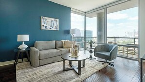 A modern living room with a blue accent wall, a grey sofa, a round coffee table, a side table with a lamp, and a view of the city through large windows. - Landing | Beautiful 1BD, Gym, Pool (Houston)