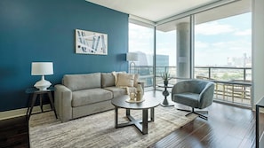A modern living room with a blue accent wall, a grey sofa, a round coffee table, a side table with a lamp, and a view of the city through large windows. - Landing | Amazing 1BD, Courtyard, Gym (Houston)