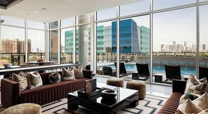 A modern hotel room with large windows, a seating area with sofas and ottomans, and a view of a city skyline and river. - Landing | Beautiful 1BD, Gym, Pool (Houston)