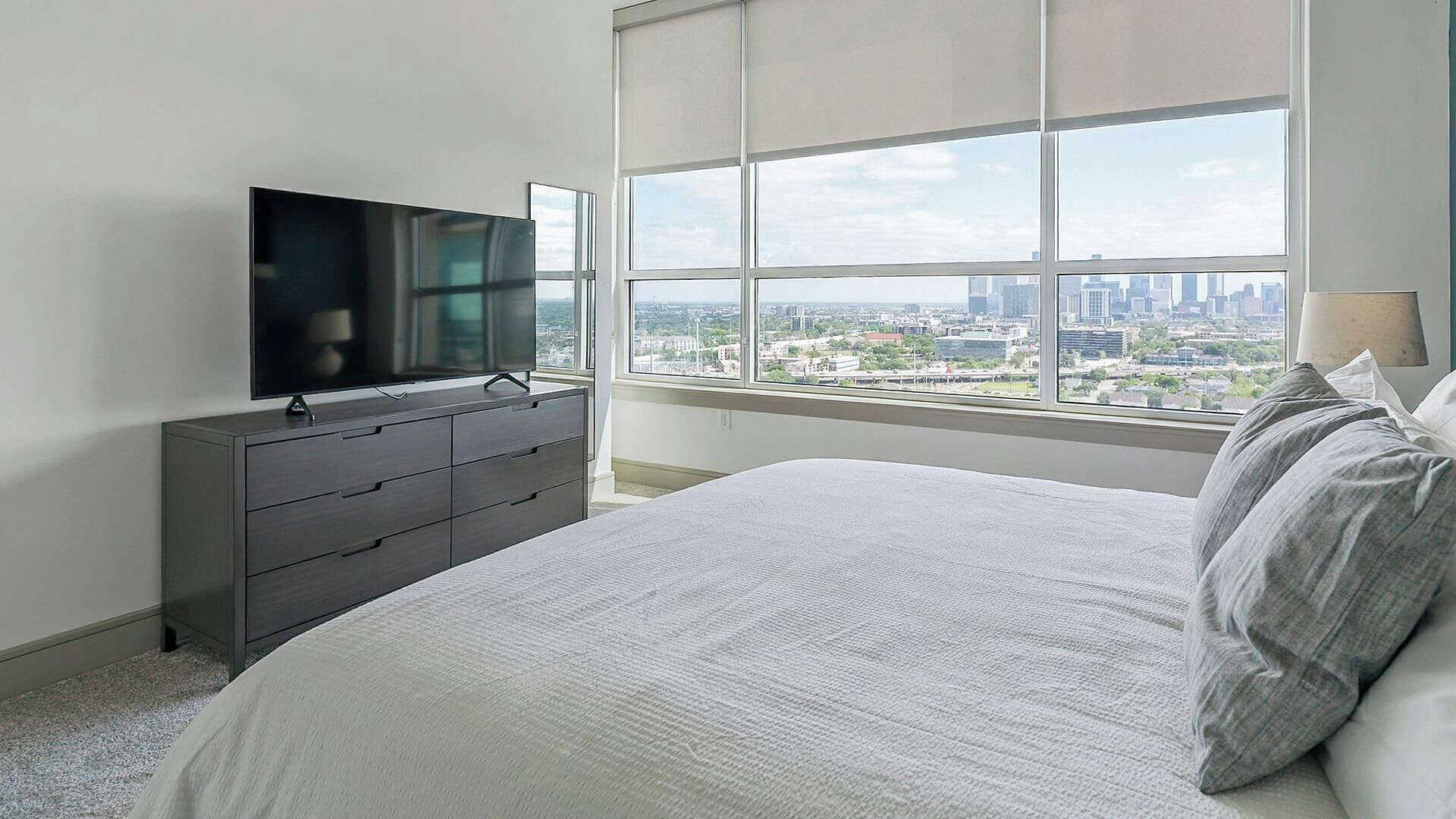 Une chambre avec un grand lit, un téléviseur posé sur une commode et une vue sur la ville grâce à de grandes fenêtres.