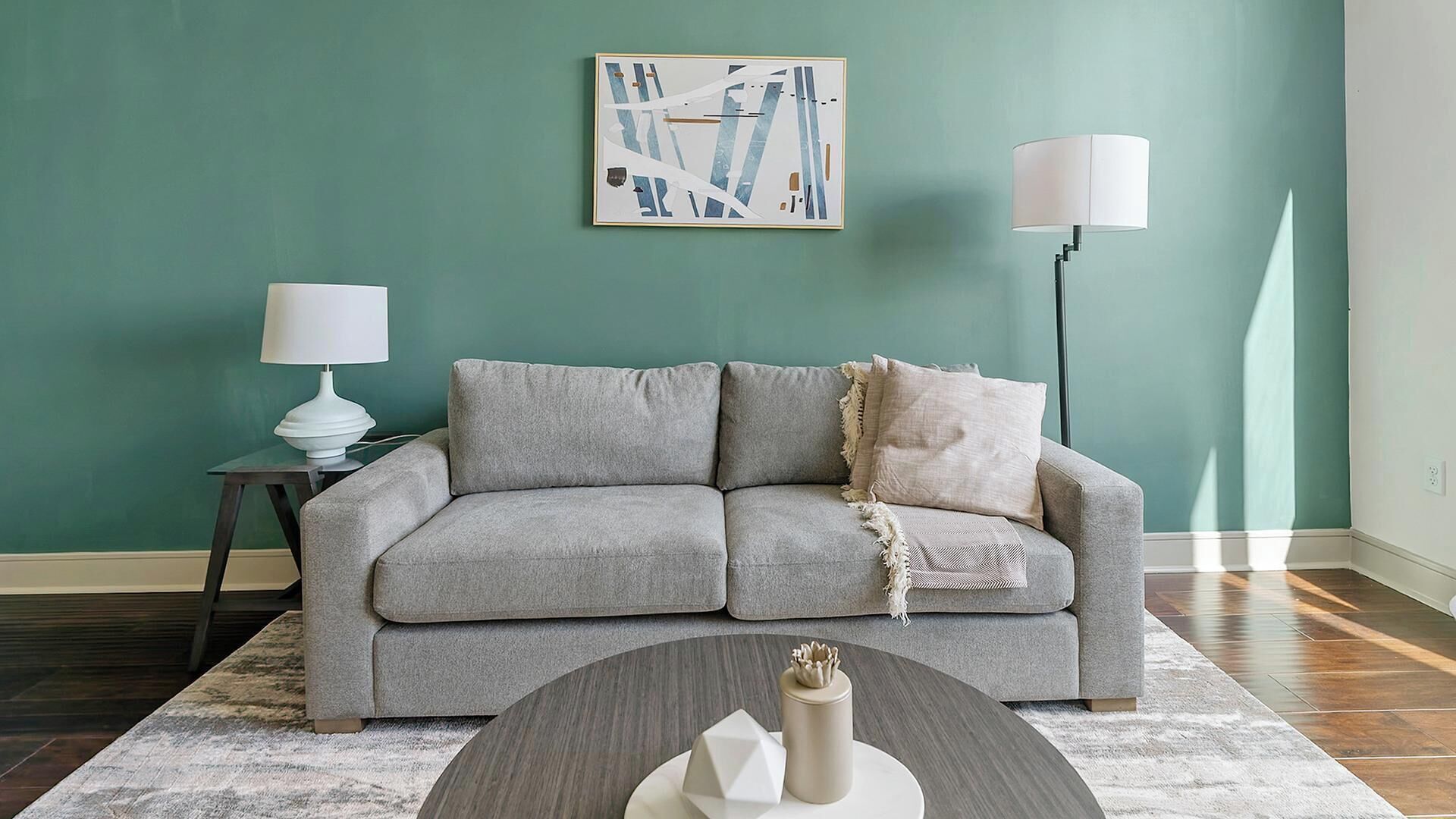 Uma sala de estar com um sofá cinza, uma mesinha lateral com uma luminária, um objeto decorativo na mesa de centro e um quadro na parede.