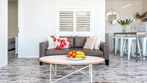 Uma sala de estar com um sofá marrom, uma mesa de centro redonda com frutas, uma parede branca e uma janela com persianas.