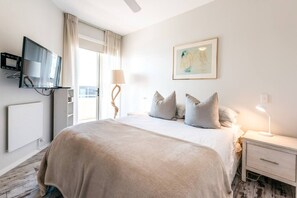 Une chambre à coucher avec un lit, un téléviseur fixé au mur, une lampe de chevet, un tableau encadré et une fenêtre avec des rideaux.
