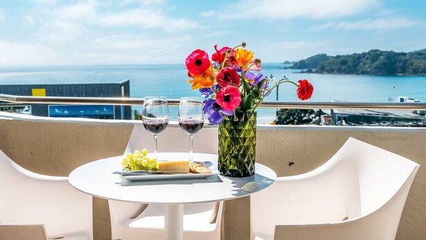 Uma mesa com um vaso de flores, dois copos de vinho tinto e um prato com queijo e uvas em uma varanda com vista para um corpo de água.