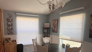 Een eetgedeelte met een ronde tafel en witte stoelen, grote ramen met zonwering en decoratieve muurkunst.