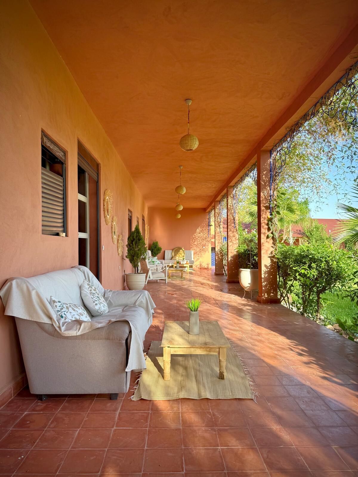 A covered outdoor seating area with a sofa and a small table, surrounded by potted plants and a view of greenery.