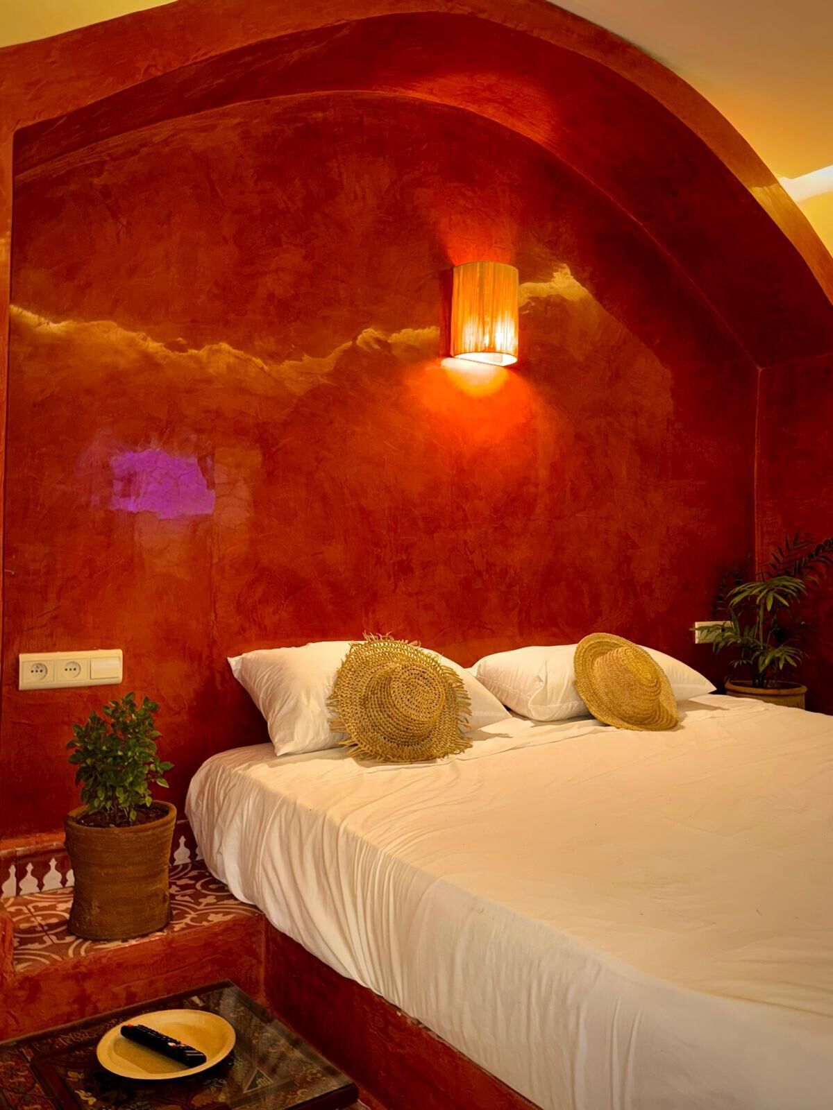 Quarto com cabeceira de parede vermelha, luminária de cabeceira, planta em vaso e controle remoto sobre a mesinha de cabeceira.