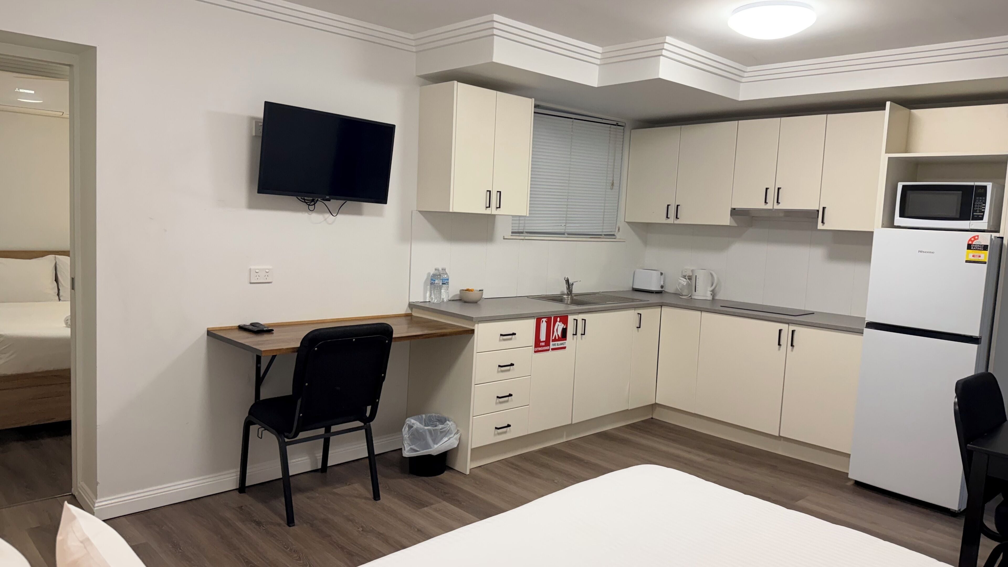 A compact hotel room with a kitchenette, a bed, a desk, and a TV.