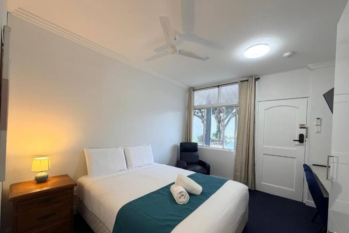 Une chambre d’hôtel équipée d’un lit, d’un bureau, d’une chaise, d’un ventilateur de plafond et d’une fenêtre avec des rideaux.