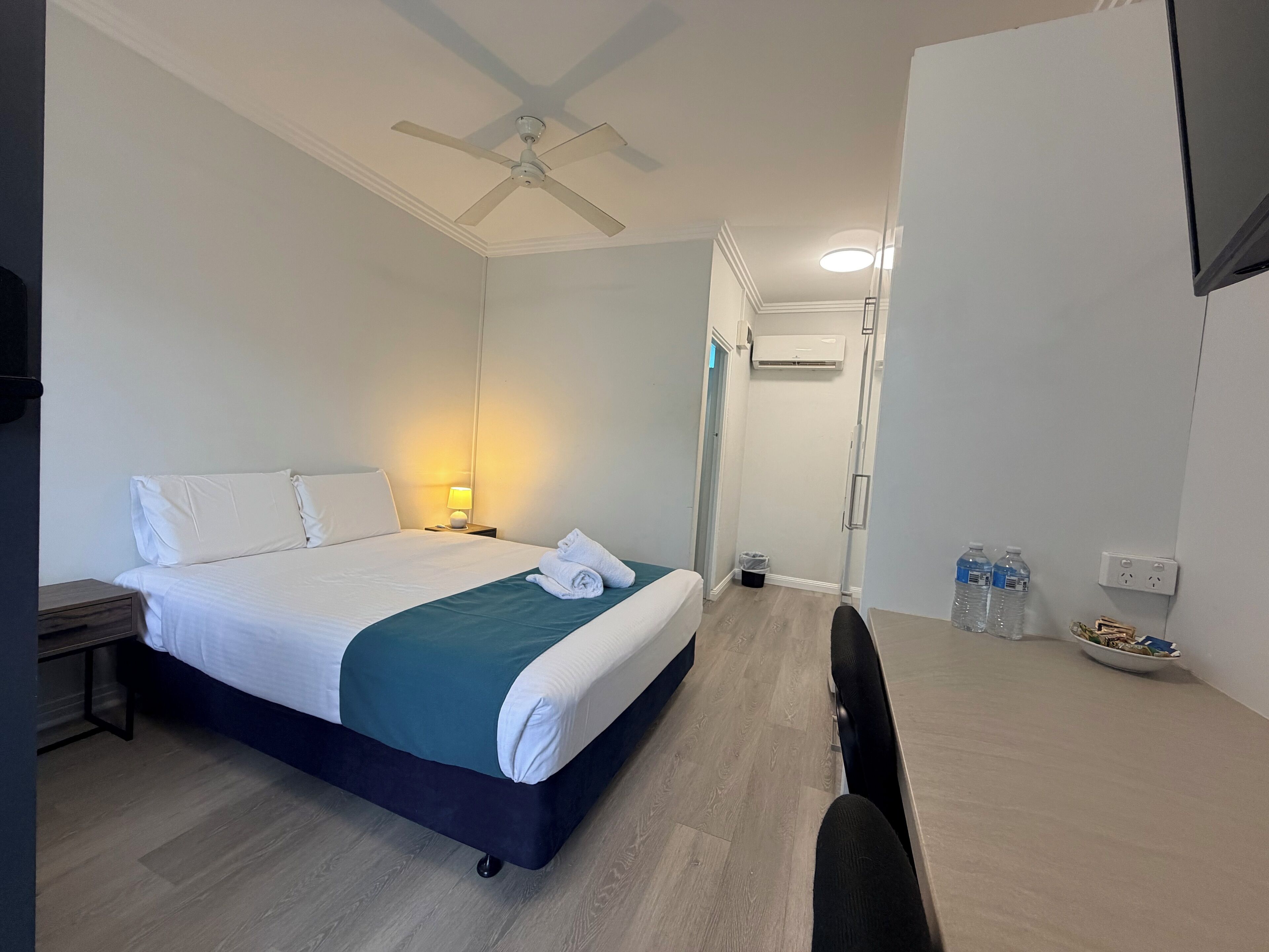 A hotel room with a bed, bedside table, lamp, ceiling fan, air conditioning unit, and a desk with a chair and water bottles.
