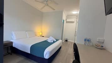 A hotel room with a bed, bedside table, lamp, ceiling fan, air conditioning unit, and a desk with a chair and water bottles.