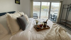 Un dormitorio con una cama, una bandeja con comida y bebidas, un balcón con sillas y vistas al exterior.