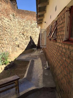 A narrow alleyway with a stone wall on one side and a brick wall on the other. There is a bench in the foreground and a window with a metal frame on the brick wall.