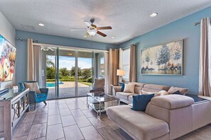 Ein geräumiges Wohnzimmer mit einer großen Couch, einem Couchtisch und einem Fernseher. Eine Deckenlampe, ein Bild an der Wand und Blick auf einen Pool im Freien sind ebenfalls vorhanden.