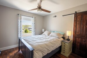 A bedroom with a bed, a ceiling fan, a nightstand with a lamp, a wooden door, and a window with curtains. - The Lookout at Buckleberry View, EV Charging (Spring Mills)