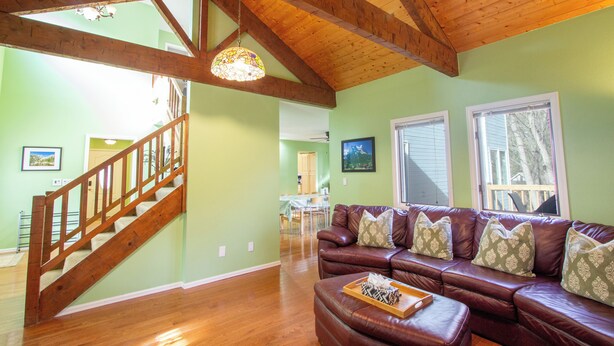 Uma sala de estar ampla com escada de madeira, sofá de couro com almofadas estampadas e um espaço de jantar ao fundo.