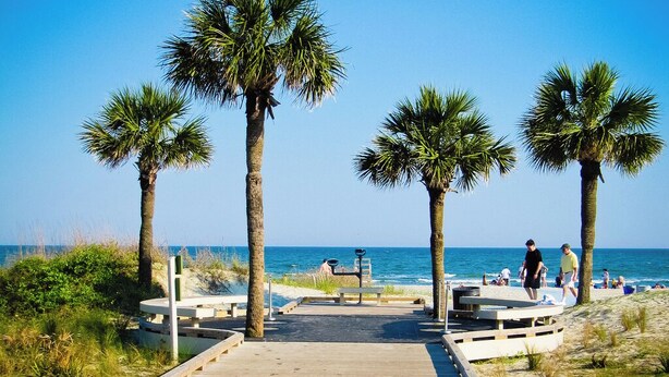 A wooden boardwalk leading to a beach with palm trees and benches. - Island Links - 3BR/2BA - Great Pool Complex! (Hilton Head Island)