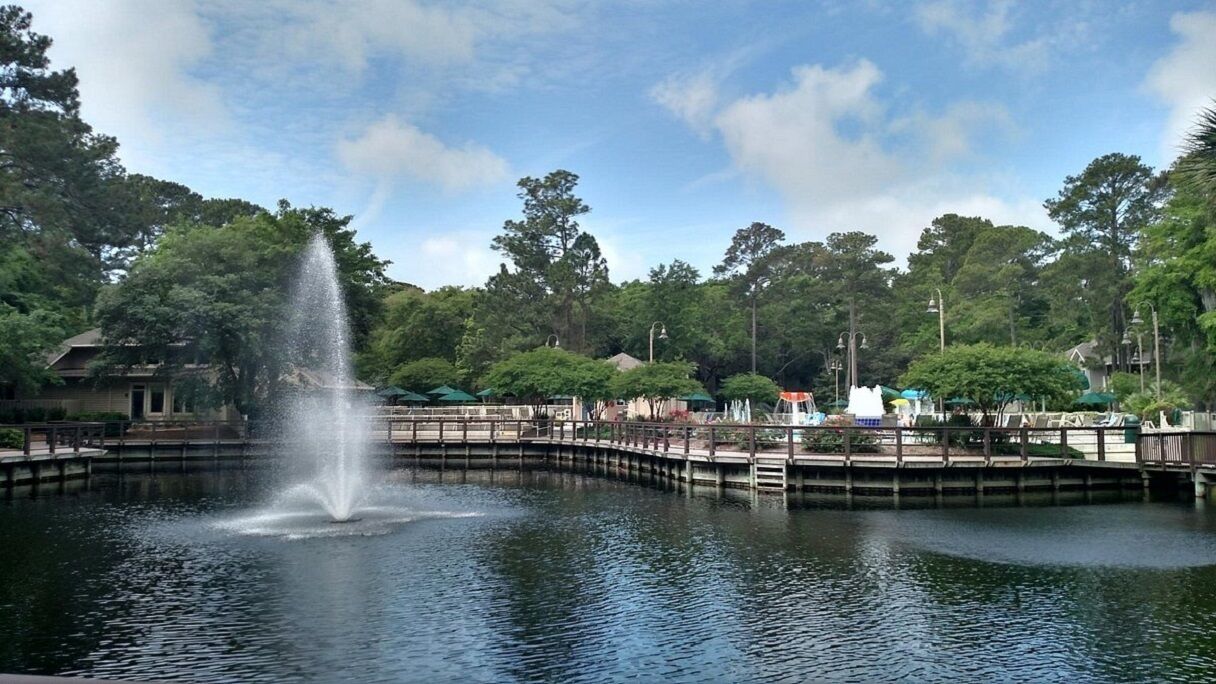 A tranquil park with a central fountain, surrounded by lush greenery and a wooden bridge.