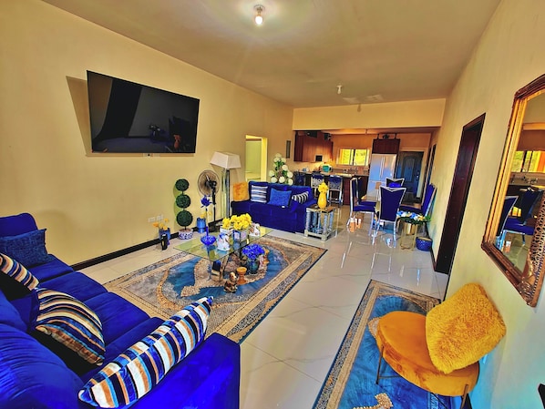 A living room with a sofa set, a coffee table with decorative items, a flat-screen TV mounted on the wall, and a kitchen area in the background.