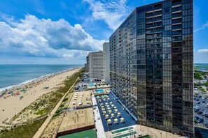 Een hoog hotel met uitzicht op het strand, meerdere balkons en een zwembadgedeelte met parasols.