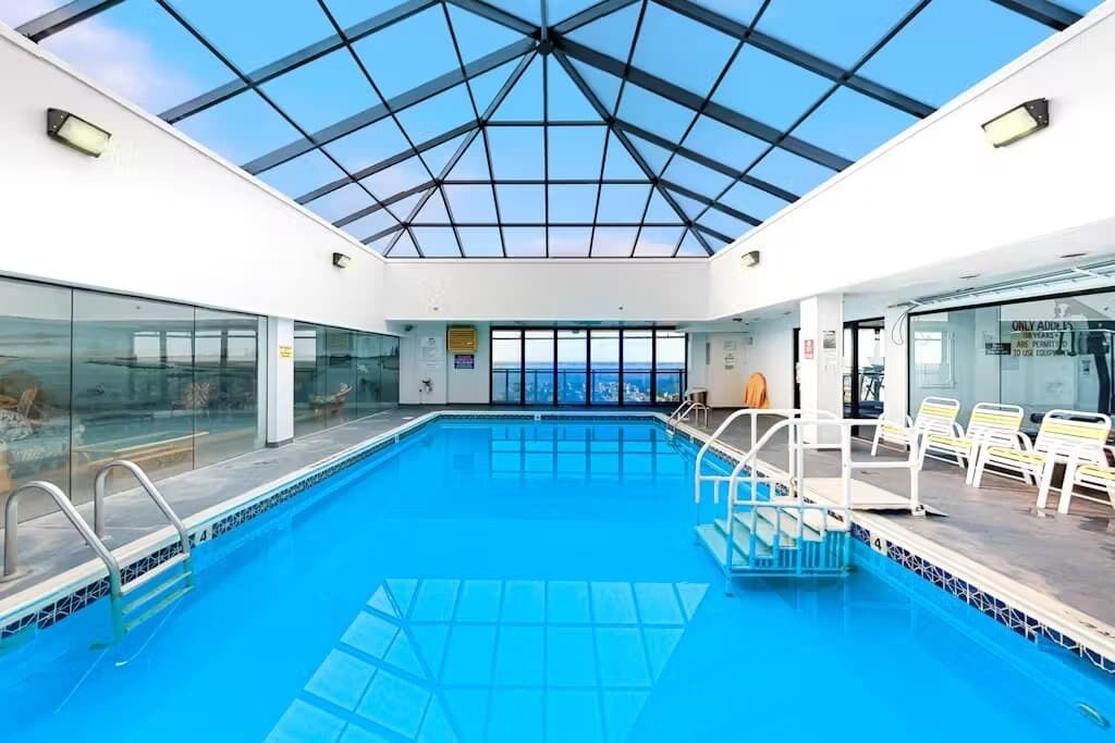 Uma piscina interior com teto de vidro, vista clara para o mar e área da piscina com espreguiçadeiras.