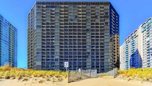 A high-rise building with a grid-like facade, surrounded by sand dunes and other buildings. - Golden Sands 1407 (Ocean City)
