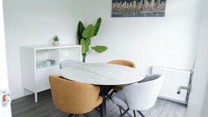 A dining area with a round marble table, two chairs, a white cabinet with glass doors, a large green plant, and a cityscape painting on the wall. - Abe’s Home (London)