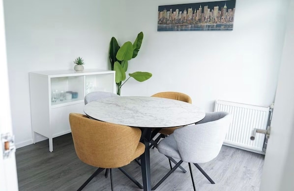 A dining area with a round marble table, two chairs, a white cabinet with glass doors, a large green plant, and a cityscape painting on the wall.