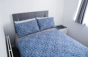 A bed with a patterned blue and white duvet, a grey headboard, and a bedside table with a black object on it.