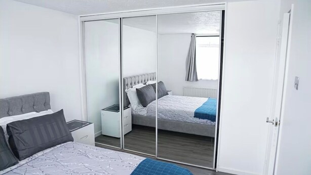 A bedroom with a large sliding closet, a bed with a grey headboard, and a bedside table with a lamp. - Abe’s Home (London)