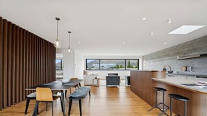 A modern kitchen with a wooden accent wall, a dining area with a wooden bench, and a bar with stools. - Rubus Street Residence | Brand-New 4 Bedroom, 4 Bathroom Retreat in Wanaka (Wanaka)