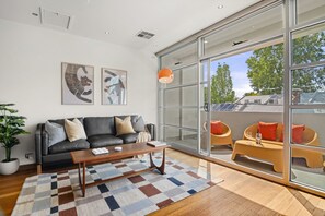 Een moderne woonkamer met een zwarte leren bank, een houten salontafel en een vloerkleed met een patroon. Twee ingelijste kunstwerken hangen aan de muur en een grote schuifdeur leidt naar een terras met oranje stoelen en een tafel.