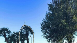 Une structure en pierre surmontée d’une croix, entourée de plantes et de fleurs en pot, située dans un espace aménagé avec des arbres et un ciel dégagé.