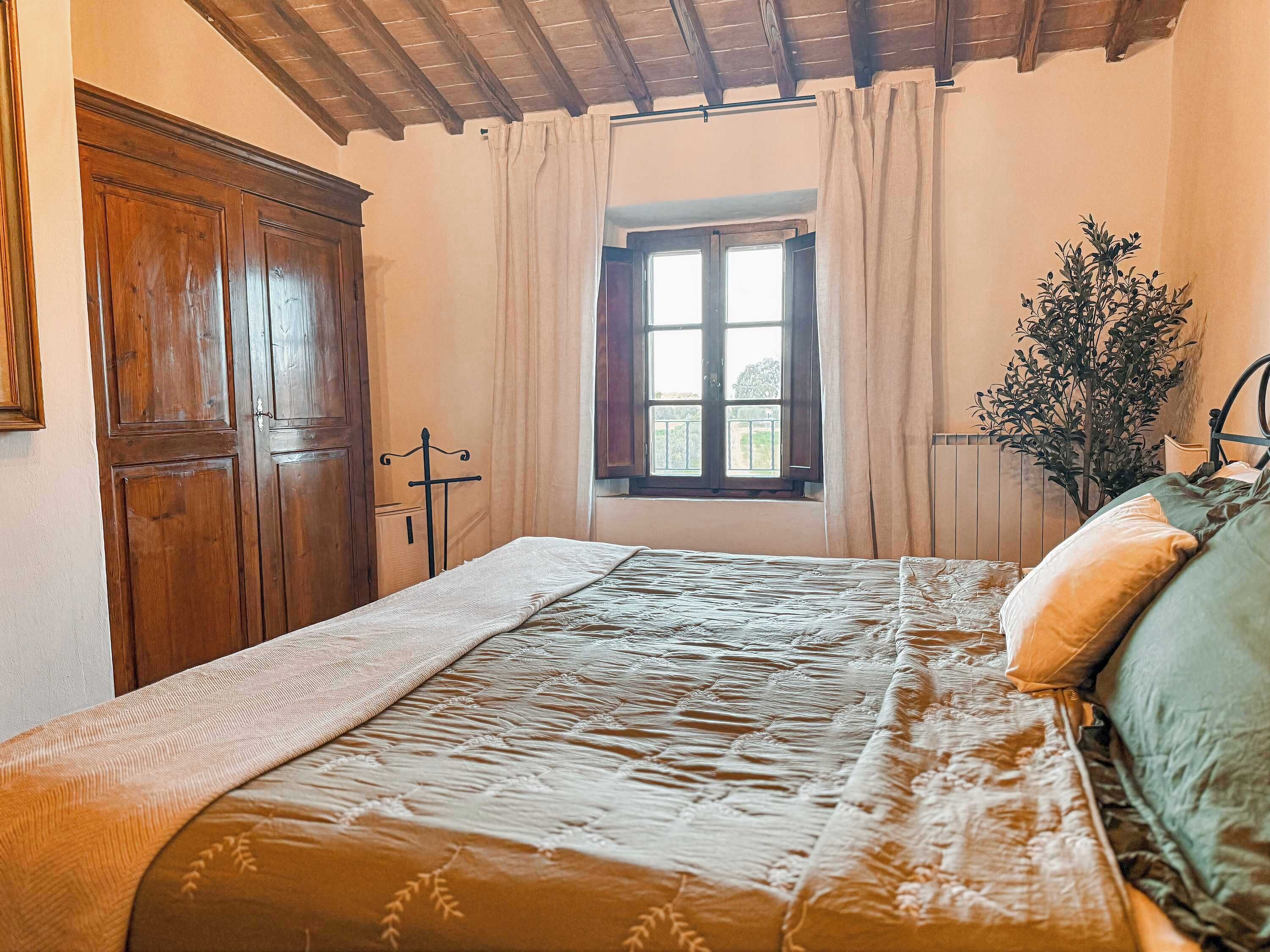 Quarto com guarda-roupa de madeira, cama com colcha estampada, janela com cortinas e um vaso de planta.