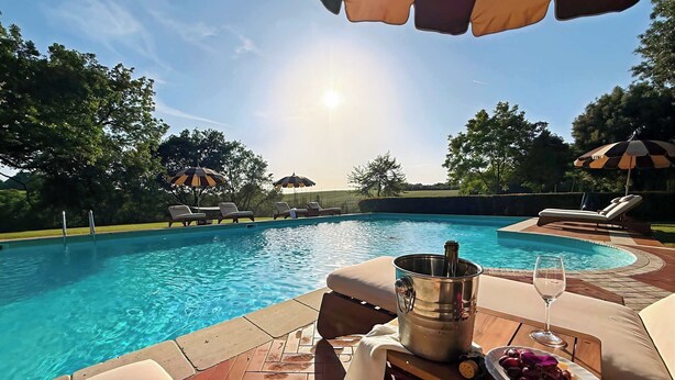 Un espace aménagé au bord de la piscine, avec des chaises longues, une table sur laquelle se trouvent une bouteille de champagne, un verre de vin et une assiette de raisins.