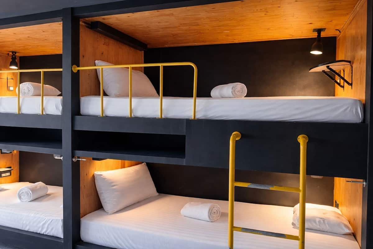 A bunk bed room with three levels, each with a mattress and pillows, and a small table with a lamp.