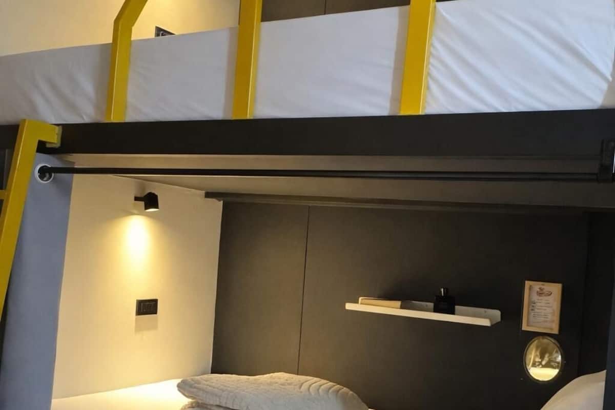 A bunk bed with a white mattress, a folded white towel, a small shelf with a framed picture, and a wall-mounted light.