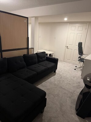 A modern living room with a sectional sofa, a small white table, a black office chair, and a closed door. - Basement with own washroom and parking (Oshawa)