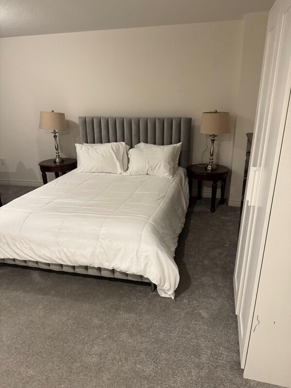 A neatly made bed with a grey upholstered headboard, two bedside tables with lamps, and a grey carpeted floor. - Basement with own washroom and parking (Oshawa)