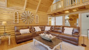 L’intérieur d’une cabane en rondins présente un grand canapé en cuir marron en forme de L, une table basse en bois et une décoration rustique comprenant un élément décoratif mural en forme de soleil rayonnant.