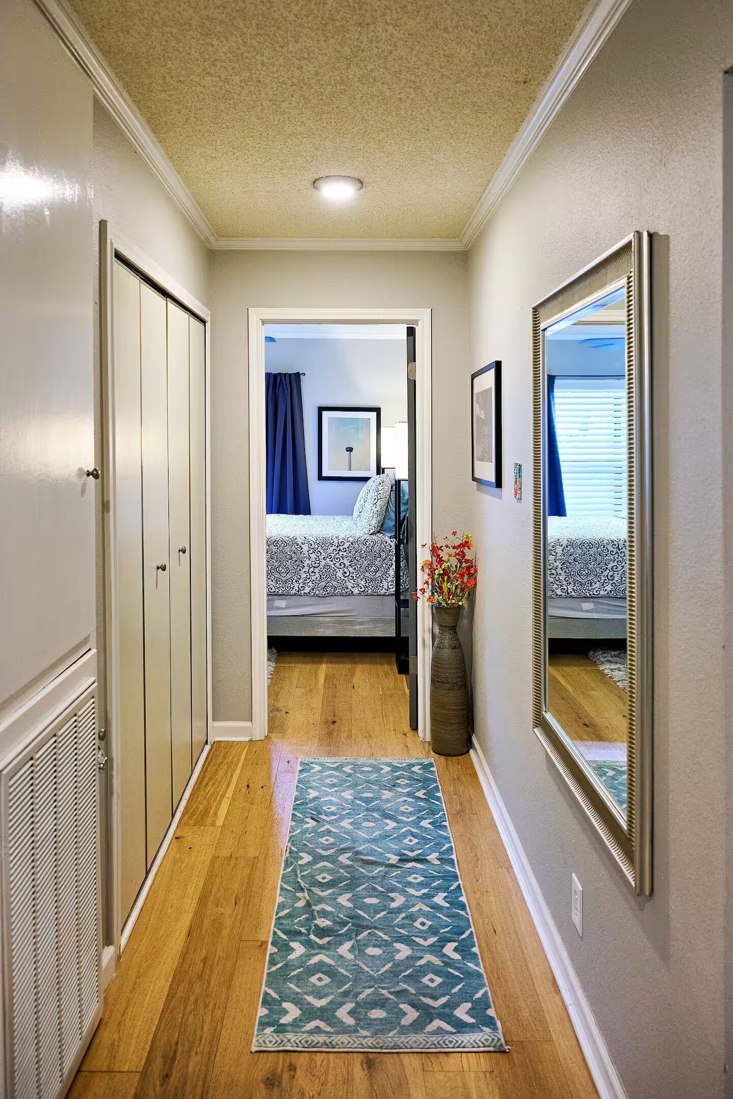 A hallway with a wooden floor, a patterned rug, a mirror, and a door leading to a bedroom with a bed and a window.