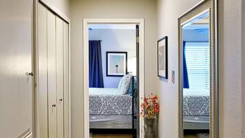 A hallway with a wooden floor, a patterned rug, a mirror, and a door leading to a bedroom with a bed and a window.