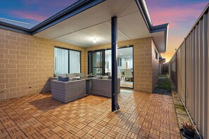 Un patio techado al aire libre con mobiliario de mimbre, piso de ladrillo y vista hacia el interior de una vivienda.