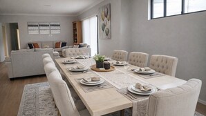 Un comedor con una mesa larga dispuesta para seis personas, rodeada por seis sillas, con un sofá y una estantería al fondo.