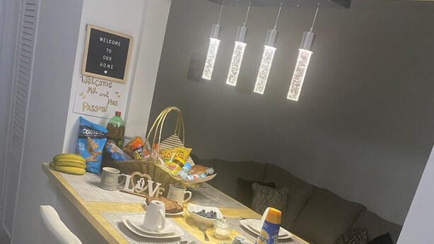 Un coin repas avec une longue table dressée pour quatre personnes, des chaises blanches, un panneau fixé au mur et des luminaires suspendus.