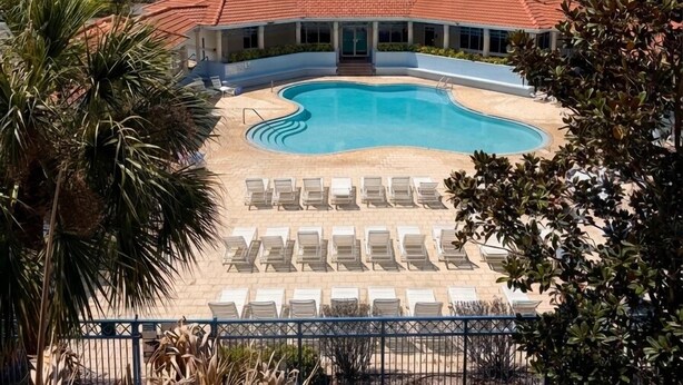 A swimming pool with sun loungers, surrounded by a fence, and modern buildings in the background. - Ocean Walk Condo: Classy Beachy Retreat in charming New Smyrna Beach (New Smyrna Beach)