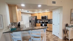 A kitchen with a central island, wooden cabinets, stainless steel appliances, and a white door. - Ocean Walk Condo: Classy Beachy Retreat in charming New Smyrna Beach (New Smyrna Beach)